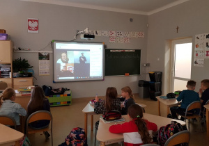 Na zdjęciu dzieci siedzące w ławkach, wpatrzone w ekran tablicy multimedialnej, na której widać autora Grzegorza Kasdepke oraz prowadzących relację.